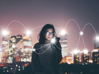 A woman in front of a cityscape at night looking at her smartphone